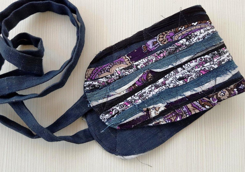 May include: A handmade fabric belt with a dark blue denim base and a colorful patchwork design featuring purple, pink, white, and brown fabric strips. The belt is tied with a long, dark blue fabric tie.