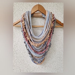 May include: A multi-layered infinity scarf in shades of gray, pink, and tan, displayed on a wooden hanger. The scarf features a mix of textures, including ruffled and smooth fabric strips, creating a unique and stylish accessory.
