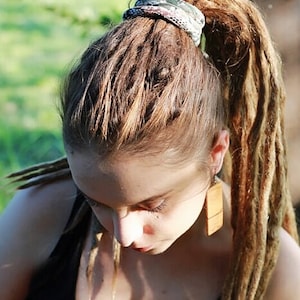 May include: A person with long, brown dreadlocks pulled back into a high ponytail, secured with a patterned scrunchie. The person is wearing a black tank top and wooden earrings. The background is blurred green.