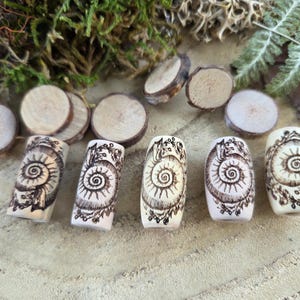 May include: Five cylindrical beads made of light-colored material, each featuring a dark brown pyrography design of a spiral ammonite fossil. The beads are arranged in a horizontal row, accompanied by several small, circular wooden slices.