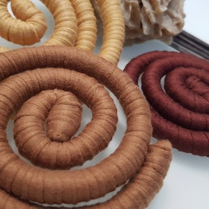 May include: Three spiral-shaped objects wrapped in fabric, featuring a light tan spiral, a medium brown spiral, and a dark burgundy spiral. Each spiral is constructed from a continuous, coiled, fabric-covered cord, creating a textured, circular form.