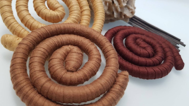May include: Three spiral-shaped objects wrapped in fabric, featuring a light tan spiral, a medium brown spiral, and a dark burgundy spiral. Each spiral is constructed from a continuous, coiled, fabric-covered cord, creating a textured, circular form.