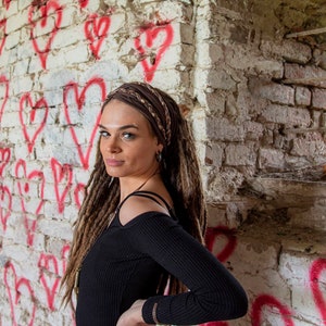 May include: A black long-sleeved ribbed top with cold-shoulder cutouts and a brown braided fabric headband worn over long brown dreadlocks. The garment features a fitted silhouette with a round neckline and long sleeves, providing a simple look.