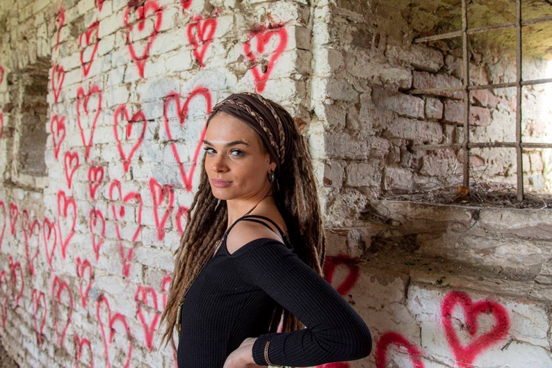 May include: A black long-sleeved ribbed top with cold-shoulder cutouts and a brown braided fabric headband worn over long brown dreadlocks. The garment features a fitted silhouette with a round neckline and long sleeves, providing a simple look.