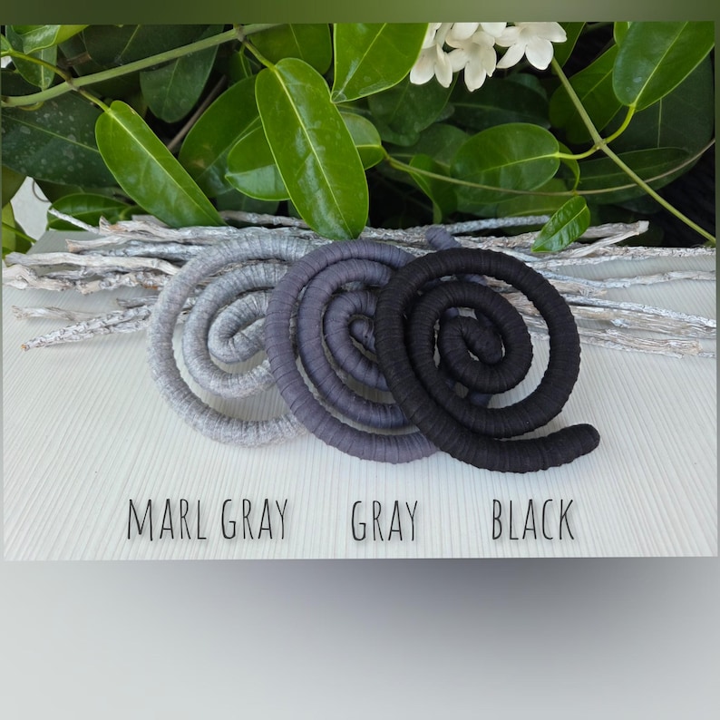 Puede incluir: Tres bobinas en forma de espiral envueltas en tela, dispuestas horizontalmente. Los colores est&aacute;n etiquetados como MARL GRAY, GRAY y BLACK. Las bobinas est&aacute;n colocadas frente a ramas blancas y hojas verdes. El texto est&aacute; impreso debajo.
