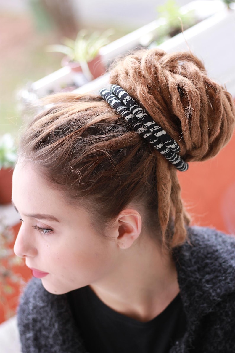 K&ouml;nnte beinhalten: Ein schwarz-wei&szlig; gemustertes Stoff-Haarband, das um einen gro&szlig;en Dutt aus hellbraunen Dreadlocks gewickelt ist. Das Haaraccessoire zeigt ein sich wiederholendes abstraktes Design und ist fest um die Basis des dicken, strukturierten Haarknotens gebunden.
