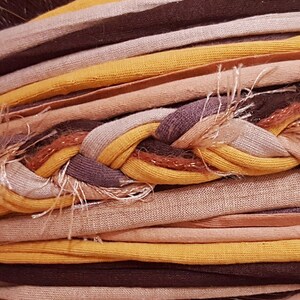 Puede incluir: Una diadema de tela de m&uacute;ltiples hilos con una secci&oacute;n central trenzada compuesta por tiras de jersey amarillo mostaza, marr&oacute;n oscuro y beige claro. El accesorio incluye bandas de tela paralelas adicionales en tonos tierra a juego con bordes deshilachados.