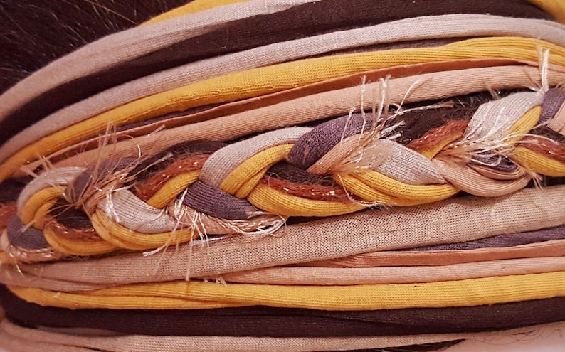Puede incluir: Una diadema de tela de m&uacute;ltiples hilos con una secci&oacute;n central trenzada compuesta por tiras de jersey amarillo mostaza, marr&oacute;n oscuro y beige claro. El accesorio incluye bandas de tela paralelas adicionales en tonos tierra a juego con bordes deshilachados.