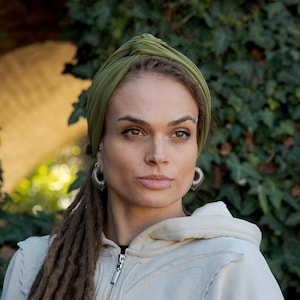 May include: An olive green fabric headband with a central knot detail worn on the head. The headband is made of a smooth, solid-colored material that wraps around the crown, providing a secure fit while keeping hair pulled back from the face.