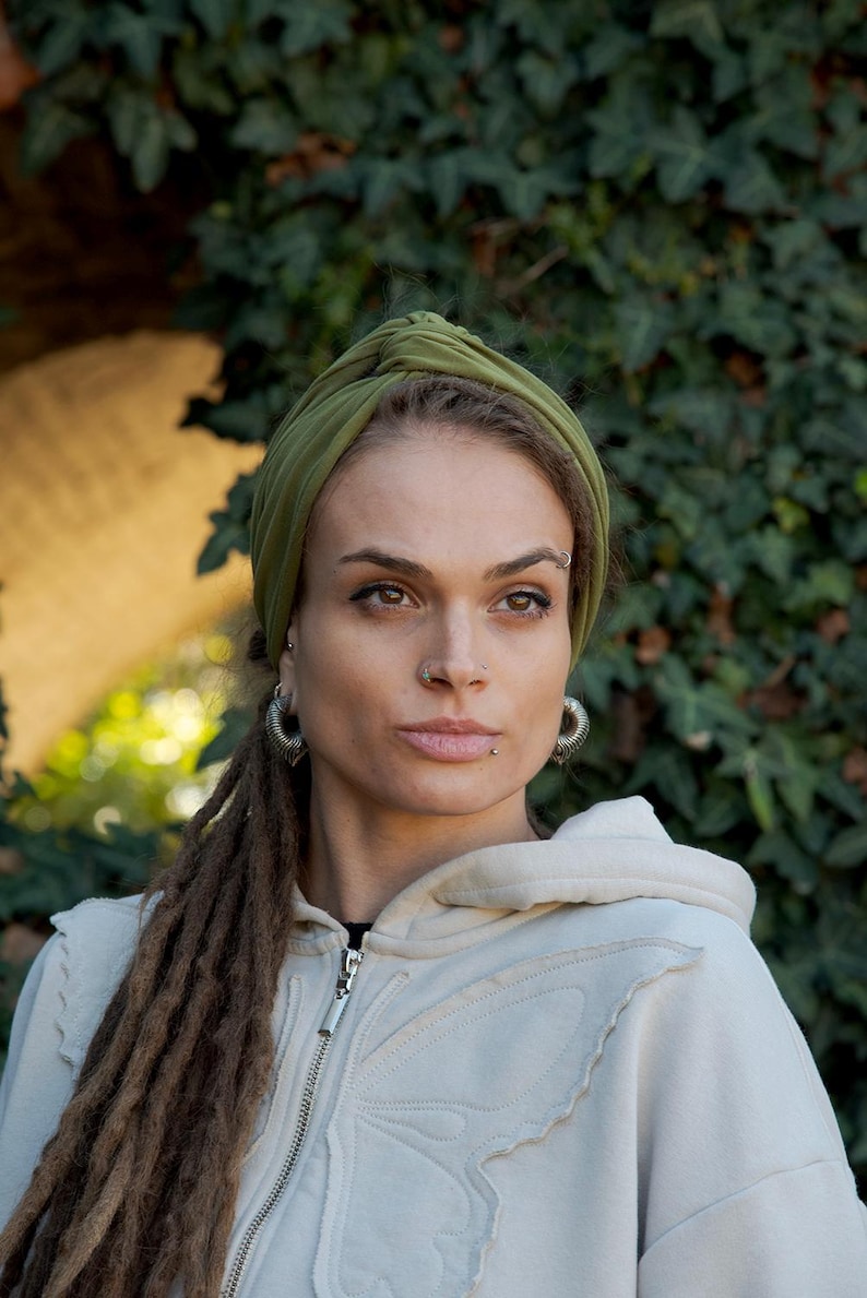 May include: An olive green fabric headband with a central knot detail worn on the head. The headband is made of a smooth, solid-colored material that wraps around the crown, providing a secure fit while keeping hair pulled back from the face.
