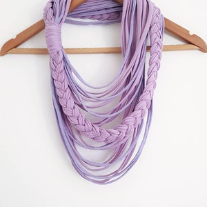 May include: A handmade lavender infinity scarf with a braided design. The scarf is made of soft fabric strips and hangs on a wooden hanger against a white background. The scarf's design features a braided section and multiple cascading strands.