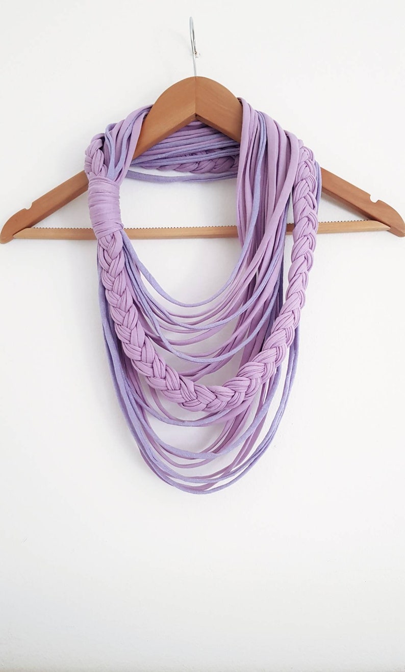 May include: A handmade lavender infinity scarf with a braided design. The scarf is made of soft fabric strips and hangs on a wooden hanger against a white background. The scarf's design features a braided section and multiple cascading strands.