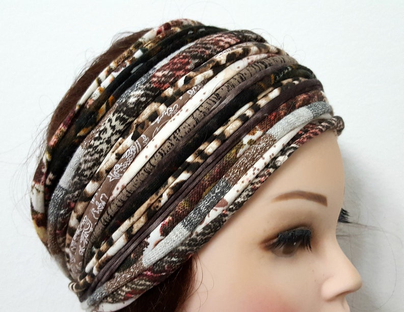 May include: A wide headband constructed from multiple strips of fabric in various brown, black, white, and tan patterns. The design features a mix of animal prints, plaid, and solid textures layered horizontally to create a thick, gathered appearance.