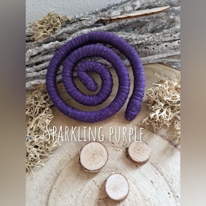 Pu&ograve; includere: Un oggetto a forma di spirale avvolto in un tessuto viola scintillante, posizionato sopra tre piccole fette di legno circolari. Il testo "SPARKLING PURPLE" &egrave; stampato in lettere bianche al centro dell'immagine, tra la spirale e il legno.