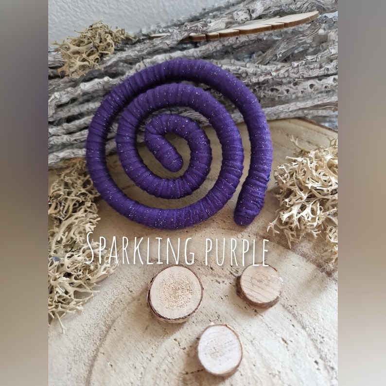 Pu&ograve; includere: Un oggetto a forma di spirale avvolto in un tessuto viola scintillante, posizionato sopra tre piccole fette di legno circolari. Il testo "SPARKLING PURPLE" &egrave; stampato in lettere bianche al centro dell'immagine, tra la spirale e il legno.