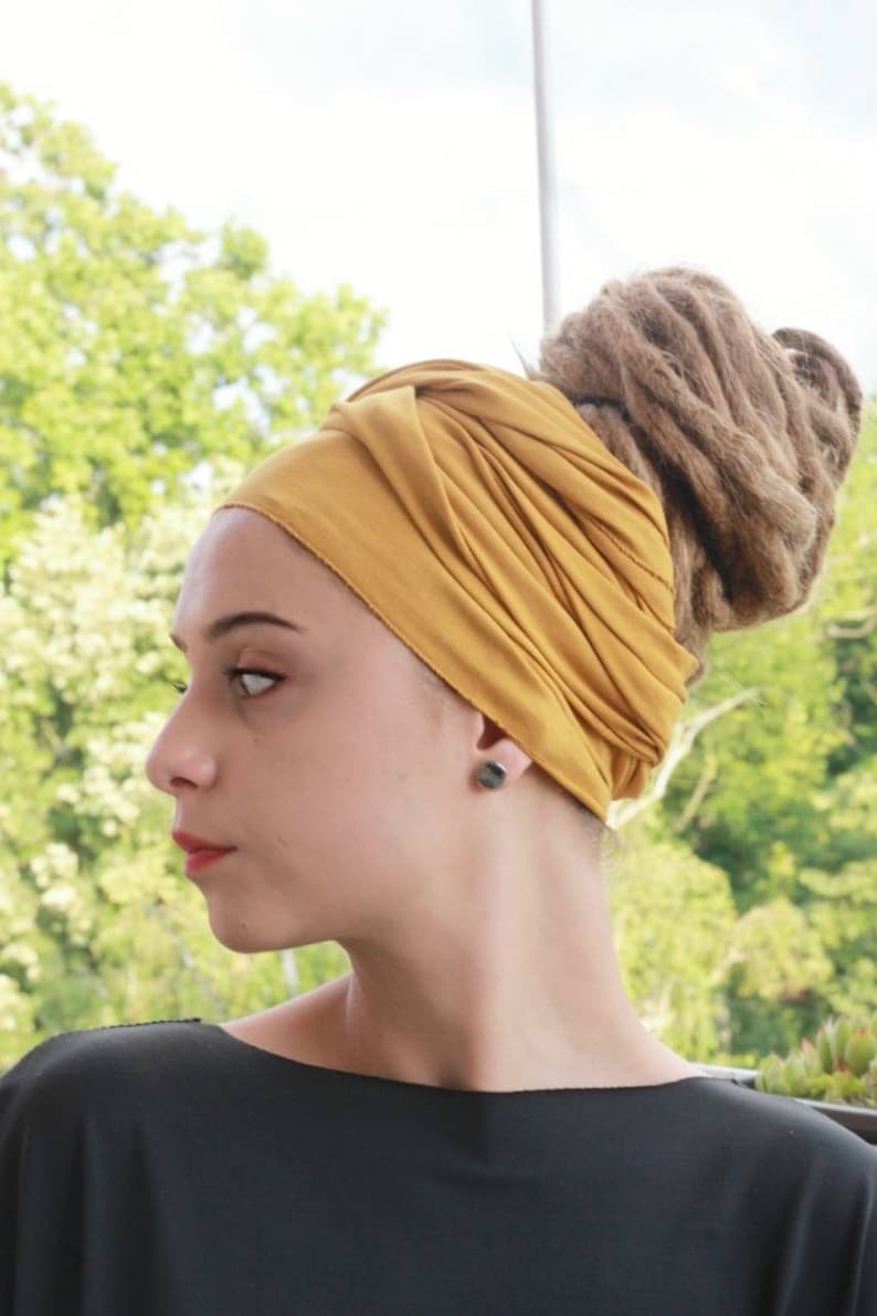 May include: A woman wearing a mustard yellow head wrap. The head wrap is wrapped around the head and covers the hair. The woman is wearing a black top and has a pair of black earrings. The background is blurred with green foliage.