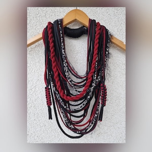 May include: A multi-layered necklace made of red and black fabric cords. The necklace is displayed on a wooden hanger against a white wall.