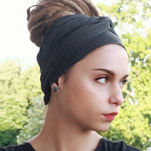 May include: A dark gray turban-style head wrap. The fabric is wrapped around the head, covering the hair. The background is a blurred view of green foliage. The person is wearing a black shirt and a dark gray earring.