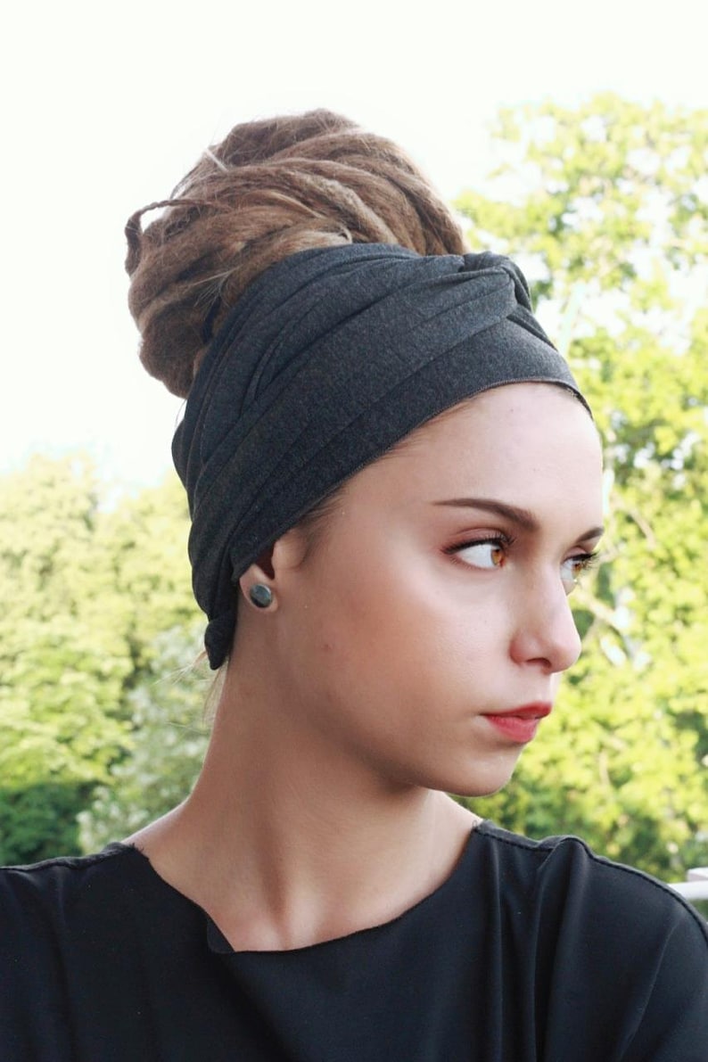 May include: A dark gray turban-style head wrap. The fabric is wrapped around the head, covering the hair. The background is a blurred view of green foliage. The person is wearing a black shirt and a dark gray earring.