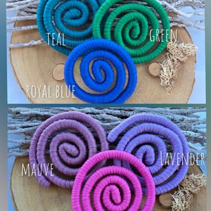 Puede incluir: Seis objetos decorativos en forma de espiral envueltos en tela, dispuestos en dos grupos. El grupo superior presenta espirales en color verde azulado, verde y azul real. El grupo inferior presenta espirales en color malva, lavanda y rosa.