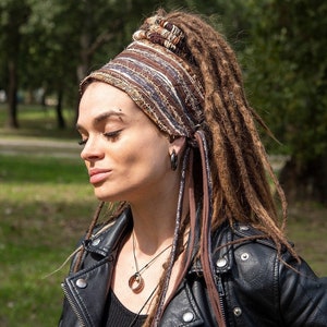 Könnte beinhalten: Eine Frau mit langen braunen Dreadlocks trägt ein braunes und weiß gestreiftes Stirnband mit langen braunen und weißen Bändern. Sie trägt eine schwarze Lederjacke und eine silberne Halskette mit einem runden Anhänger.
