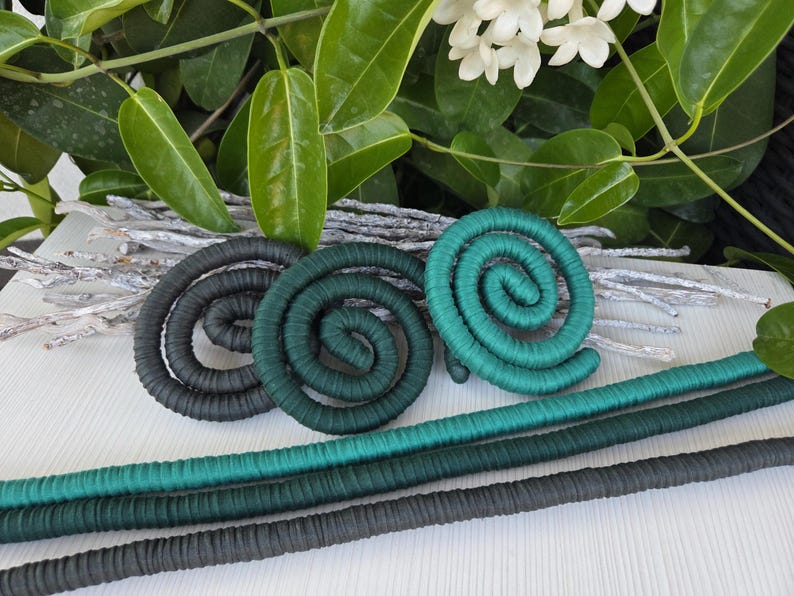 Puede incluir: Tres adornos en forma de espiral envueltos en tela, con un degradado de colores desde el gris carb&oacute;n oscuro hasta el verde bosque profundo y el azul cerceta brillante. Debajo de las espirales hay tres varillas largas y rectas de tela.