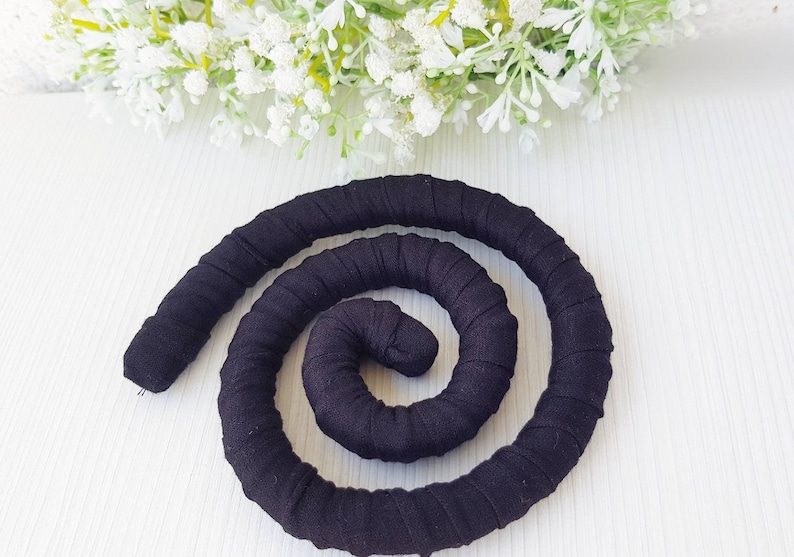 May include: A flexible, black fabric-covered coil shaped into a spiral. The material is wrapped tightly around a cylindrical core, creating a continuous, textured, and segmented appearance throughout the entire length of the dark, solid-colored object.
