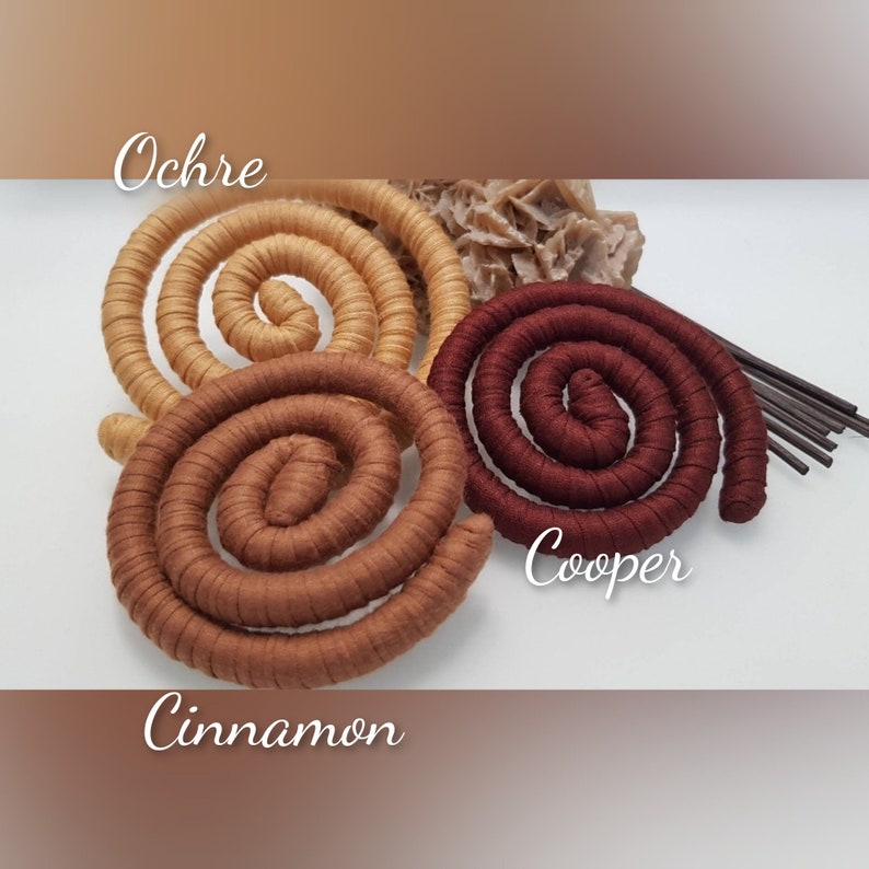 May include: Three spiral-shaped decorative objects wrapped in fabric, labeled Ochre, Cinnamon, and Cooper. The spirals are arranged in a cluster with a small, textured, light-colored mineral formation and several thin, dark brown sticks visible behind them.