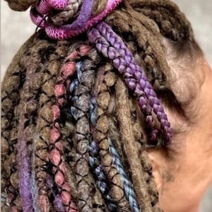 Pu&ograve; includere: Un insieme di dreadlock castani acconciati in uno chignon alto, arricchiti da ciocche sintetiche nei toni vivaci del viola, blu e rosa. I capelli presentano intrecci complessi e un aspetto testurizzato e annodato con vari riflessi colorati.