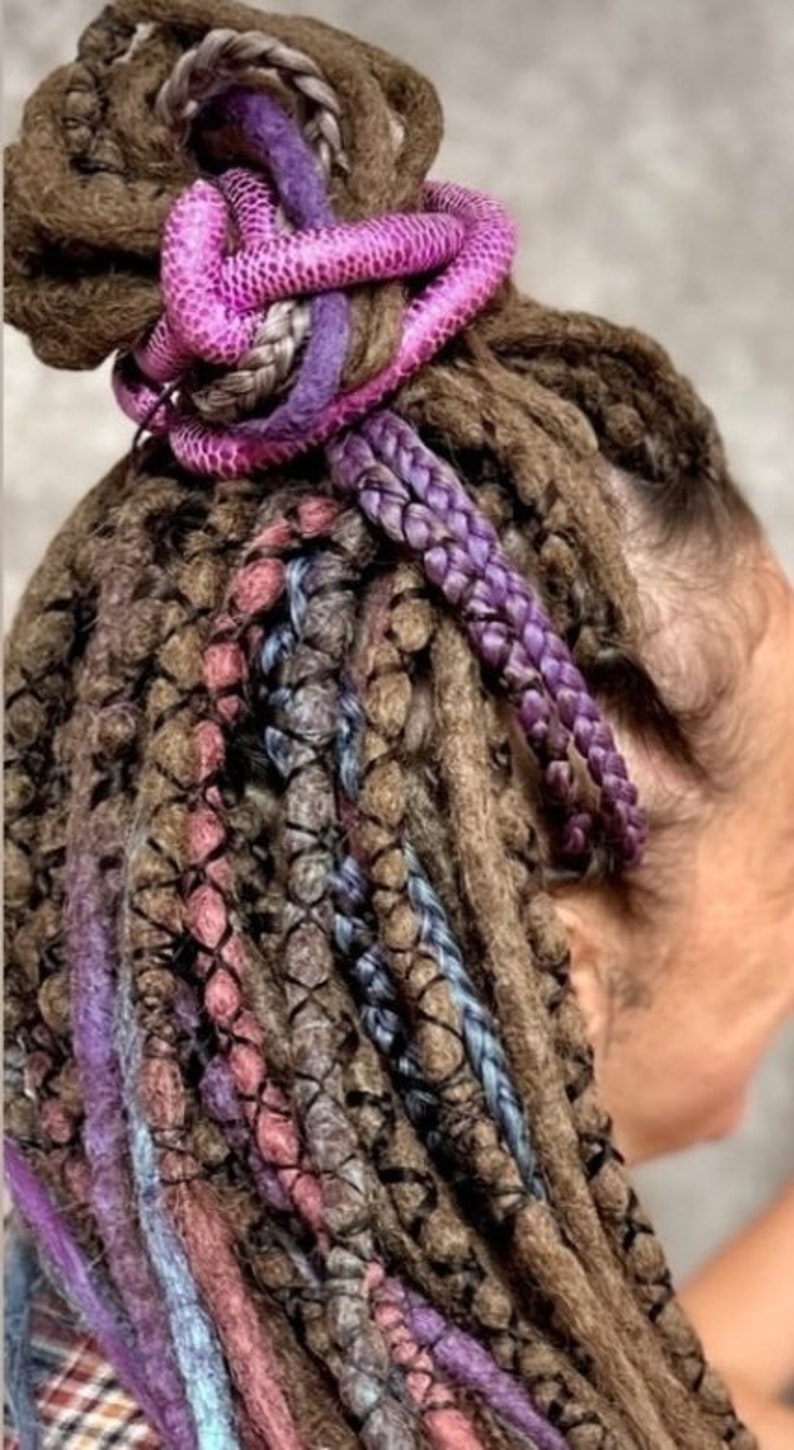 Pu&ograve; includere: Un insieme di dreadlock castani acconciati in uno chignon alto, arricchiti da ciocche sintetiche nei toni vivaci del viola, blu e rosa. I capelli presentano intrecci complessi e un aspetto testurizzato e annodato con vari riflessi colorati.
