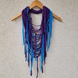 May include: A purple and blue crocheted necklace with fringe hanging from the bottom. The necklace is hanging on a wooden hanger.