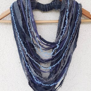 May include: A multi-strand infinity scarf constructed from various strips of denim and fabric in shades of dark blue, grey, and light blue. The design features frayed edges and a textured, layered appearance, with a central band holding the strands.