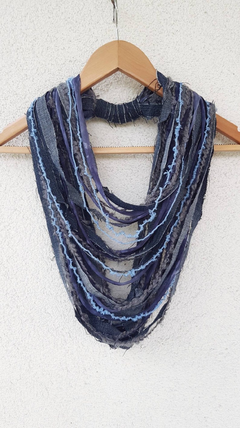May include: A multi-strand infinity scarf constructed from various strips of denim and fabric in shades of dark blue, grey, and light blue. The design features frayed edges and a textured, layered appearance, with a central band holding the strands.