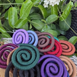 Puede incluir: Una colecci&oacute;n de adornos en forma de espiral envueltos en tela de colores, incluyendo tonos morado, azul, verde oscuro, negro, marr&oacute;n, rojo y naranja, dispuestos en un grupo superpuesto sobre una base de madera circular.