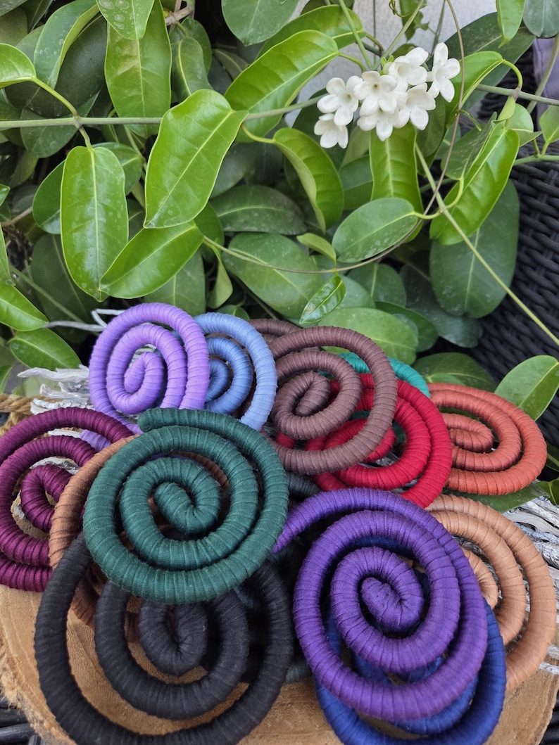 Puede incluir: Una colecci&oacute;n de adornos en forma de espiral envueltos en tela de colores, incluyendo tonos morado, azul, verde oscuro, negro, marr&oacute;n, rojo y naranja, dispuestos en un grupo superpuesto sobre una base de madera circular.