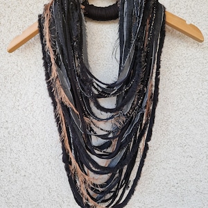 Pode incluir: Um cachecol comprido, preto e castanho, feito de tiras de tecido com bordas desfiadas. O cachecol est&aacute; pendurado num cabide de madeira.