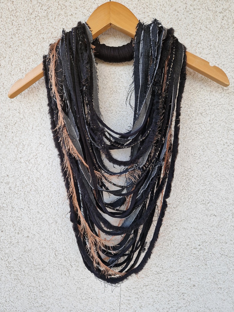 Pode incluir: Um cachecol comprido, preto e castanho, feito de tiras de tecido com bordas desfiadas. O cachecol est&aacute; pendurado num cabide de madeira.