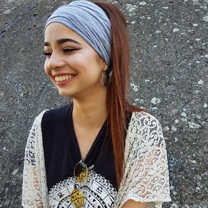 May include: A gray headband worn with a black V-neck shirt and a white lace shawl. Sunglasses are clipped to the shirt. The person is smiling, with long brown hair pulled back in a ponytail.