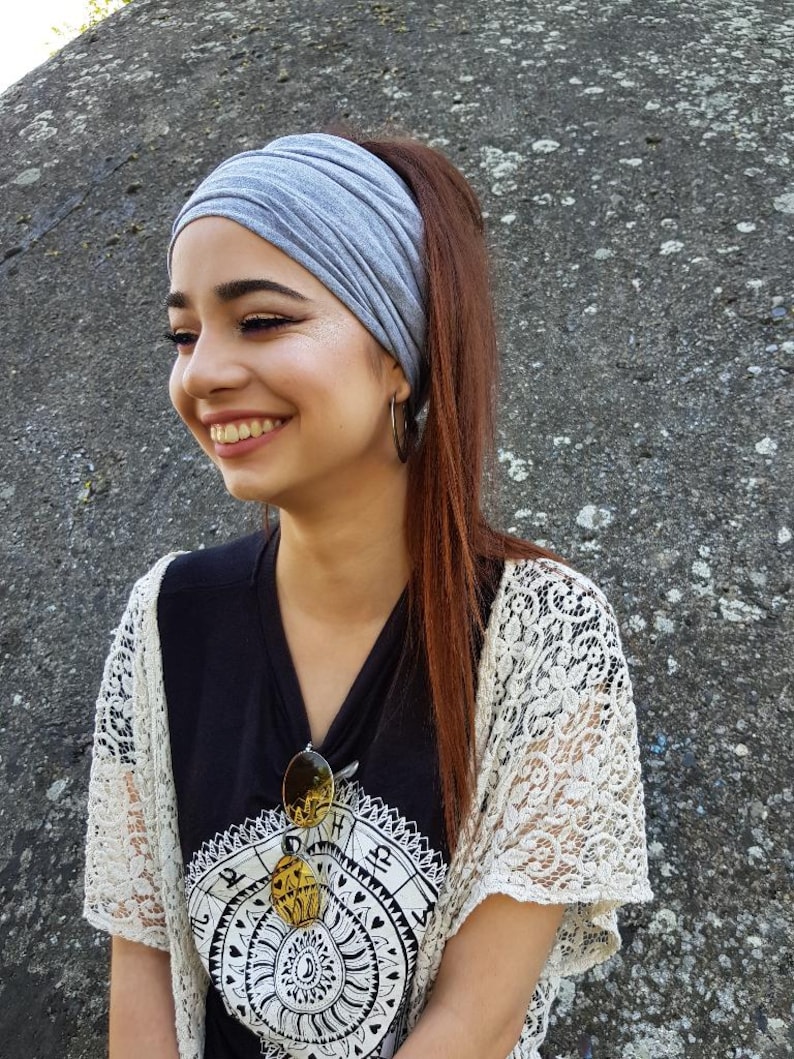 May include: A gray headband worn with a black V-neck shirt and a white lace shawl. Sunglasses are clipped to the shirt. The person is smiling, with long brown hair pulled back in a ponytail.