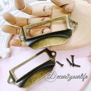 May include: Two antique brass label holders with a rectangular frame and curved bottom. The holders have a distressed finish and come with small screws for mounting. The label holders are shown on a wooden hand.