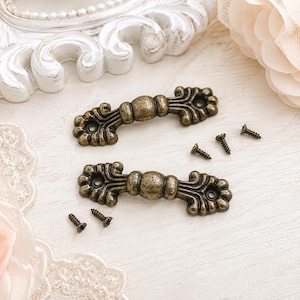 May include: Two antique brass-toned metal drawer pulls with a central rounded knob and ornate, scalloped ends featuring screw holes. The set includes four small metal mounting screws, each with a pointed tip and a threaded shank for installation.