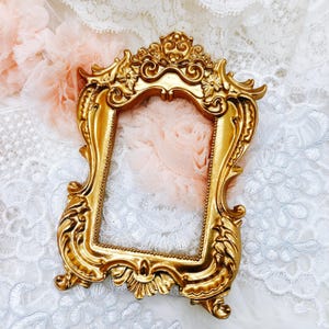 May include: Ornate gold-colored picture frame with intricate floral and scroll detailing. The frame has a rectangular opening and is set against a backdrop of white lace and pink fabric. A decorative accent for home decor.