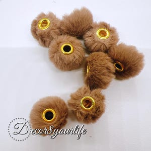 May include: Seven brown faux fur pom poms with gold metal eyelets. The pom poms are approximately 2 inches in diameter.