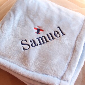 May include: A light blue baby blanket with the name "Samuel" embroidered in navy blue, along with a small red and blue airplane design. The blanket has a soft, plush texture and a stitched border.