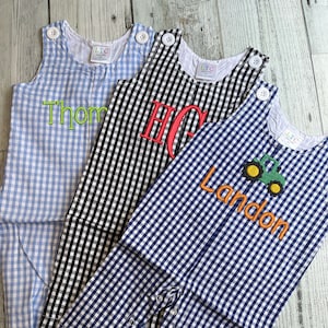 May include: Three baby rompers in blue, black, and navy gingham patterns. The blue romper has "Thom" embroidered in green. The black romper has "HC" embroidered in red. The navy romper has "Landon" and a tractor embroidered in orange and green.