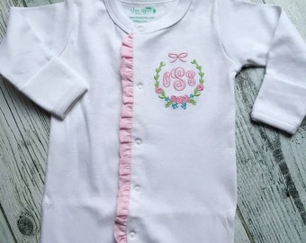 May include: White baby footed sleeper with a pink ruffle down the front and a floral monogram. The sleeper has long sleeves and snaps down the front. The monogram includes the letters "SG" inside a wreath.