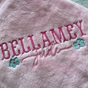 May include: A soft, pink blanket with the name "BELLAMEY" embroidered in a vibrant pink script. The name "Jude" is embroidered below in a cursive font. Two small teal and pink flower designs flank the name. The text "The Embroidered Touch" is at the bottom.