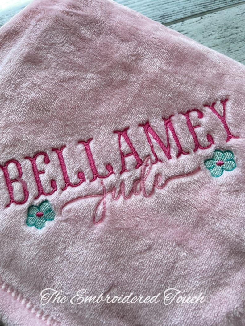 May include: A soft, pink blanket with the name "BELLAMEY" embroidered in a vibrant pink script. The name "Jude" is embroidered below in a cursive font. Two small teal and pink flower designs flank the name. The text "The Embroidered Touch" is at the bottom.
