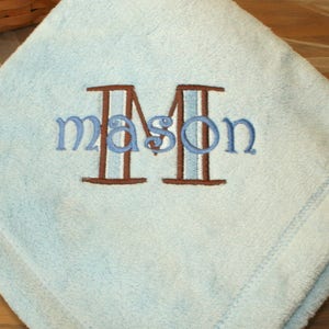 May include: A light blue baby blanket with the name "Mason" embroidered in blue thread. The letter "M" is brown with a light blue stripe. The blanket is folded and has a stitched border.