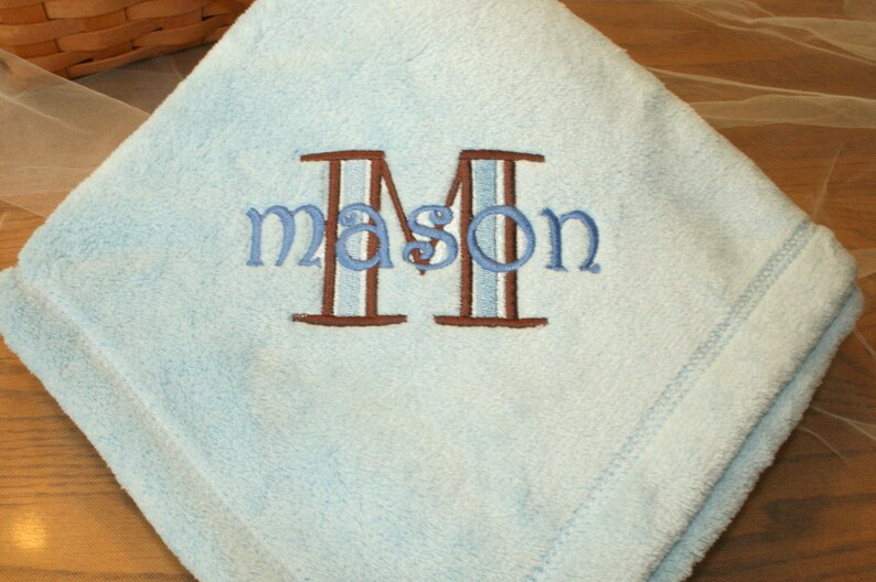 May include: A light blue baby blanket with the name "Mason" embroidered in blue thread. The letter "M" is brown with a light blue stripe. The blanket is folded and has a stitched border.